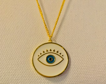 Puede incluir: Un collar de cadena dorada con un colgante circular blanco con un diseño de ojo azul malvado.