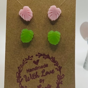 Puede incluir: Cuatro pendientes de botón con forma de hoja en dos pares, presentados en una tarjeta marrón. Dos pendientes son de color rosa claro y dos son verdes. La tarjeta tiene el texto "Handmade With Love by Lena" en una fuente decorativa.