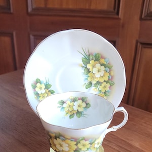Può includere: Un set vintage di tazza e piattino da tè. La tazza e il piattino in porcellana bianca presentano un motivo floreale con fiori gialli e verdi. La tazza è su un piccolo supporto dorato. Il set è posto su una superficie di legno.