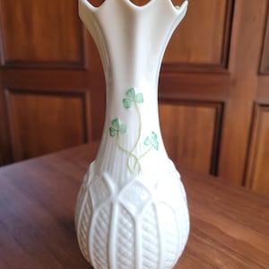 May include: Cream-colored ceramic vase with a scalloped rim and a bulbous base. The vase features a delicate green shamrock design and embossed leaf and diamond patterns. A decorative piece for home decor.