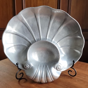 May include: A silver-colored, shell-shaped decorative dish. The dish has a textured surface with radiating lines and a central, rounded depression. It is supported by a black, wrought-iron stand, and is displayed on a wooden surface.