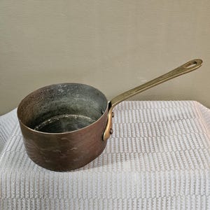 May include: A round copper saucepan with a hammered exterior finish and a long, straight brass handle attached with two rivets. The interior of the pan shows a dark, weathered metallic patina, and the handle features a circular hole at the end.