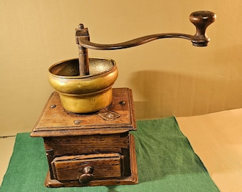 Op de afbeelding: Een antieke koffiemolen met een houten basis en een messing maalbak. De molen heeft een handslinger en een kleine lade. De houten basis is donkerbruin en de messing bak heeft een gouden tint. De molen staat op een groene doek.
