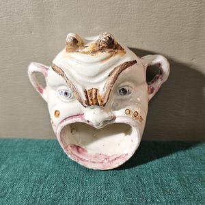 May include: A white ceramic face-shaped container with a wide open mouth. The container has two small handles and is decorated with brown and pink accents. The face has a stern expression with blue eyes and a furrowed brow.