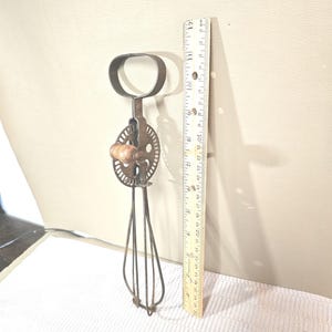 Pode incluir: Um batedor de ovos manual de metal vintage com cabo de madeira e um laço superior circular. O batedor está posicionado ao lado de uma régua de madeira, mostrando um comprimento total de aproximadamente 30 cm do laço superior até a base.