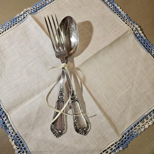 May include: A silver fork and spoon tied together with a yellow ribbon, resting on a white linen napkin with blue and white crochet trim.