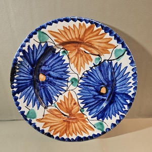May include: Round ceramic plate with a white background, featuring a floral design in blue and orange. The plate's edge has a scalloped border in blue. Green accents are also present in the floral design.