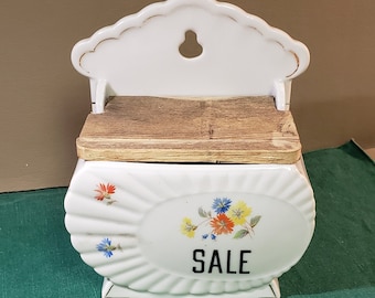 Puede incluir: Un dispensador de sal de pared de cerámica blanca con tapa de madera y diseño floral. El dispensador tiene la palabra "SALE" impresa en la parte delantera.