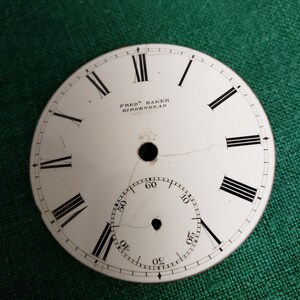 May include: White clock face with black Roman numeral hour markers and black minute markers. The clock face has a crack in it. The text "FREDK BAKER BIRKENHEAD" is printed on the clock face.