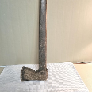 May include: An antique axe with a weathered, dark wooden handle and a metal head. The axe head is rectangular with a slightly curved blade. The handle is long and shows signs of age and use. The overall appearance suggests a tool used for chopping wood.