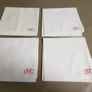 Może przedstawiać: Cztery białe lniane serwetki, każda złożona i z czerwonym haftowanym monogramem. Serwetki są starannie ułożone w stos, z monogramem widocznym na rogu każdej. Tło jest w neutralnym beżowym kolorze.