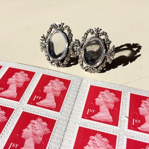 May include: Two silver oval-shaped frames with intricate filigree designs. The frames are set against a background of red and white postage stamps featuring the profile of Queen Elizabeth II.