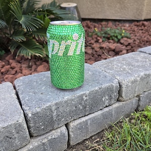 May include: A green Sprite can covered in sparkling green rhinestones. The word "Sprite" is spelled out in white rhinestones. The can is sitting on a gray stone wall.