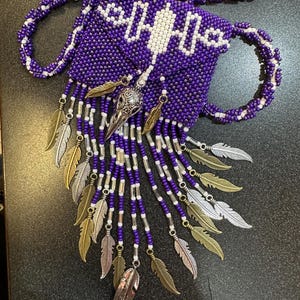 May include: A purple beaded necklace with a small beaded pouch and a silver and gold feather charm. The pouch has a white beaded design and a silver skull charm.