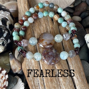 May include: Two bracelets made of agate beads in shades of green, brown, and white. The bracelets are layered on a piece of driftwood with the word "FEARLESS" printed below.