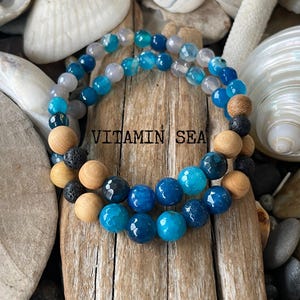 May include: Two beaded bracelets with blue agate, wood, and lava beads. The bracelets are stacked on a piece of driftwood with the text "VITAMIN SEA" visible.