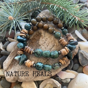 May include: Two beaded bracelets with turquoise and wooden beads. The bracelets are displayed on a piece of driftwood, with the text "NATURE HEALS" below. The background includes rocks, shells, and pine needles.