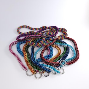 May include: A collection of colorful chainmail necklaces in various colors, including red, blue, green, black, and gold. The necklaces are made of small metal rings linked together in a pattern. The necklaces are all different lengths and have a variety of clasps.
