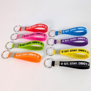 Peut inclure: Un ensemble de neuf porte-clés en silicone de couleurs rouge, rose, vert, orange, bleu, violet, jaune et noir. Chaque porte-clé possède un anneau en métal argenté et le texte imprimé en blanc SIT. STAY. OBEY. avec une icône de patte.