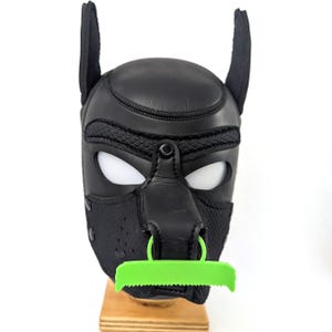 Puede incluir: Una capucha de cachorro de cuero negro con detalles de malla, inserciones blancas para los ojos y un protector bucal verde en forma de sierra. La capucha presenta orejas verticales y un anillo de metal en la nariz.