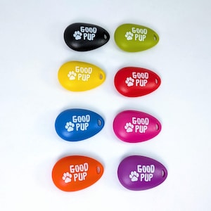 May include: A collection of eight colorful, oval-shaped pet tags. Each tag is a different color: black, green, yellow, red, blue, pink, orange, and purple. The tags have the words "GOOD PUP" and a paw print in white.