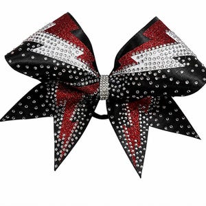 May include: A black and red cheerleading bow with a sparkling design. The bow features a red lightning bolt design with white rhinestones and a black base. The centre of the bow is adorned with a rhinestone band.