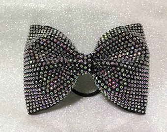 May include: Black hair bow adorned with iridescent rhinestones. The bow is a classic shape, with a central knot and two loops on either side. The rhinestones catch the light, creating a sparkling effect.