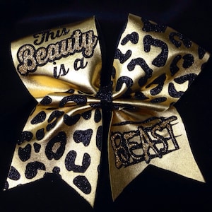 May include: Gold and black cheer bow with a leopard print design. The bow has the text "This Beauty is a Beast" written on it.
