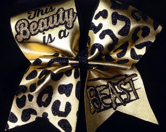 May include: Gold and black cheer bow with a leopard print design. The bow has the text "This Beauty is a Beast" written on it.
