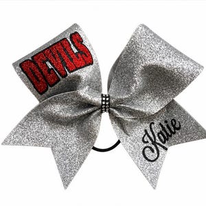 May include: Silver glitter cheerleading bow with the word "DEVILS" in red glitter and the name "Katie" in black script. The bow is secured with a black elastic band and a rhinestone centre.