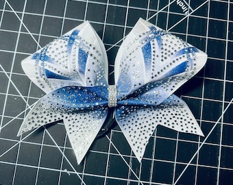 May include: A large white and blue cheer bow with silver glitter and rhinestones. The bow has a white center with a blue ombre effect on the edges.