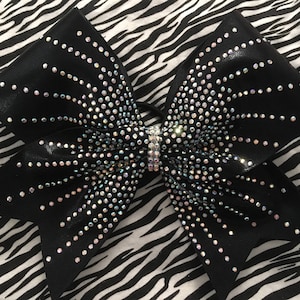 May include: A black cheerleading bow with iridescent rhinestones arranged in a radiating pattern. The bow is centered with a rhinestone-studded band. The background is a black and white zebra print.