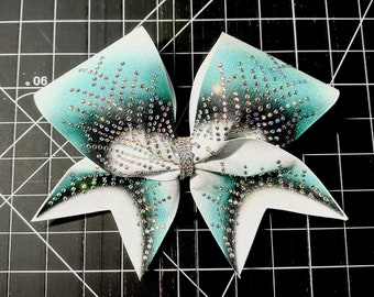 Pode incluir: Um laço de torcida branco e azul-turquesa com strass iridescentes. O laço é feito de um tecido macio e brilhante e tem uma peça central com um detalhe em metal prateado.