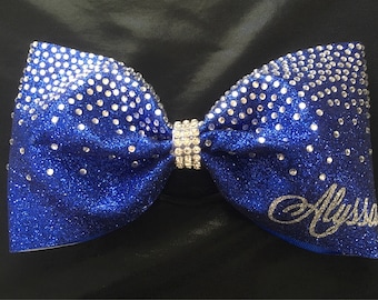 May include: A royal blue cheer bow with silver glitter and rhinestones. The bow has a silver rhinestone band in the center and the name "Alyssa" is written in silver glitter on the right side.