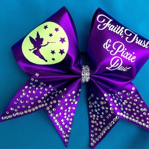 Puede incluir: Lazo de animadora morado con pedrería y un diseño de Campanilla verde que brilla en la oscuridad. El lazo tiene el texto "Faith, Trust & Pixie Dust" en un lado.