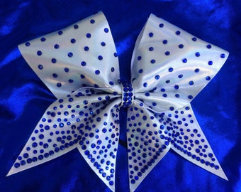 May include: A white cheer bow with blue rhinestones. The bow is made of iridescent fabric and has a pointed shape.