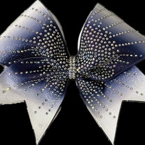 May include: A large, decorative cheerleading bow with a gradient design. The bow transitions from white to navy blue, embellished with numerous sparkling rhinestones. The center features a rhinestone-covered band, adding to its glamorous appeal.