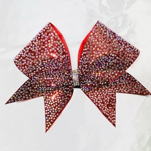 May include: A large red cheer bow with iridescent rhinestones. The bow is made of a soft, shiny fabric and has a pointed shape.