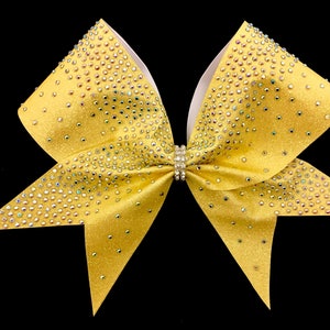 May include: A yellow cheer bow with white backing and iridescent rhinestones. The bow is tied with a silver rhinestone band.
