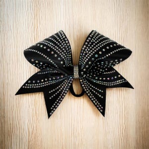 Peut inclure: Un nœud de cheerleading noir orné de strass étincelants disposés en rangées. Le nœud présente une bande centrale en strass et un élastique noir. Accessoire de cheveux décoratif.