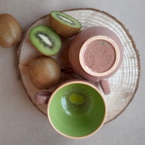 May include: Two ceramic mugs with a brown and green glaze. The mugs are on a wooden slice with kiwi fruit.