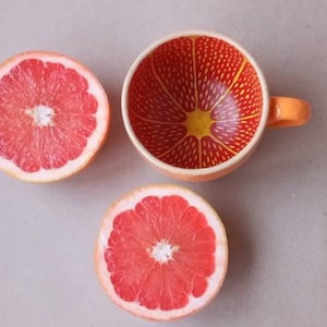 Op de afbeelding: Twee helften van een roze grapefruit en een keramische mok met een grapefruitontwerp. De mok is oranje met een geel centrum en rode lijnen.