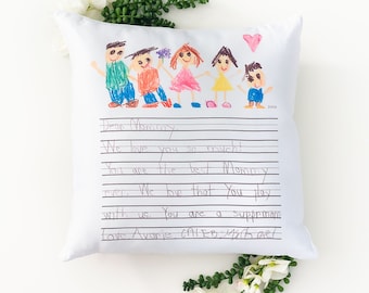 May include: A white pillow with a colorful drawing of a family on the front. The drawing is done in crayon and includes a heart. The pillow has a lined area with a handwritten message that reads "Dear Mommy, We love you so much! You are the best Mommy ever. We love that you play with us. You are a superman. Love Avarie, CALEB, Michaet".