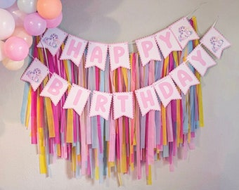 May include: A pink and white banner with the words "Happy Birthday" in pink letters. The banner is decorated with pastel colored streamers and is hanging on a white wall. There is a pink table with a rainbow colored table skirt in front of the banner.