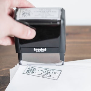 May include: A black rubber stamp with the word "trodat" printed on it. The stamp is being used to imprint a label that reads "VILLAGE CHOCOLATE CO. 21 Rider Bay Box 236, Site 200 Grand Coulee, Saskatchewan SAP 777".
