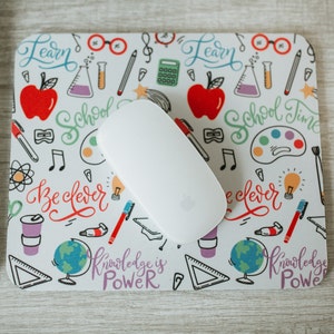 May include: A white mouse pad with a colorful school-themed design. The design includes images of pencils, paintbrushes, musical notes, and the words "Learn", "School Time", "Be Clever", and "Knowledge is Power".