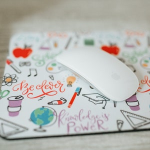 May include: A white mouse pad with a colorful doodle design featuring school supplies, a globe, and the text "Be Clever" and "Knowledge is Power".