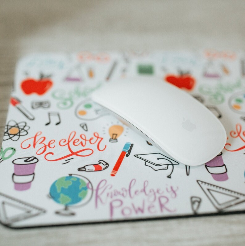 May include: A white mouse pad with a colorful doodle design featuring school supplies, a globe, and the text "Be Clever" and "Knowledge is Power".