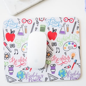 May include: White mouse pad with a colorful school-themed design. The design includes a variety of school supplies, such as pencils, rulers, and books. The text "Learn", "School Time", "Be Clever", and "Knowledge is Power" is included in the design.