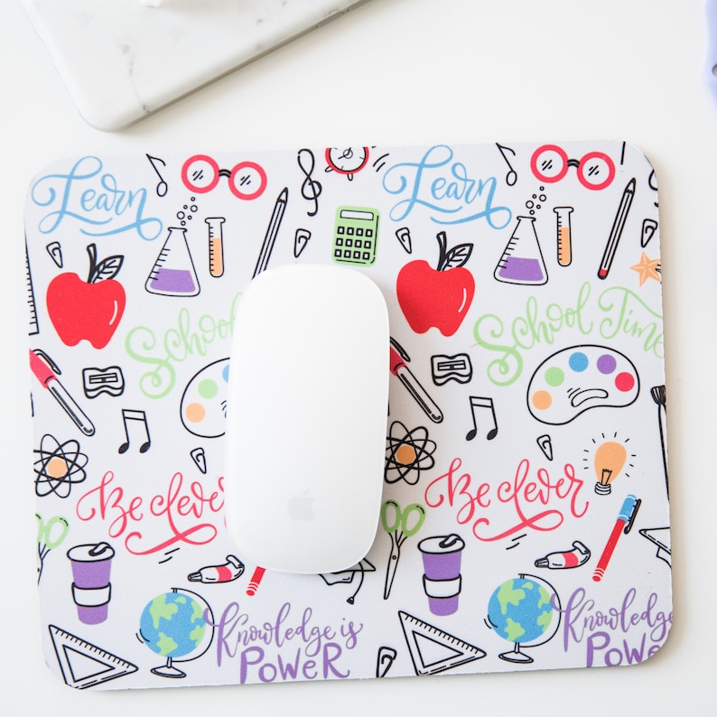May include: White mouse pad with a colorful school-themed design. The design includes a variety of school supplies, such as pencils, rulers, and books. The text "Learn", "School Time", "Be Clever", and "Knowledge is Power" is included in the design.